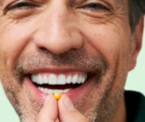 Smiling man holding a small yellow pill near his mouth.