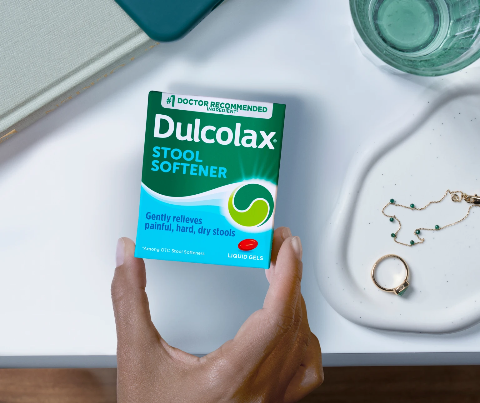 Hand holding Dulcolax Stool Softener box near a glass and jewelry.