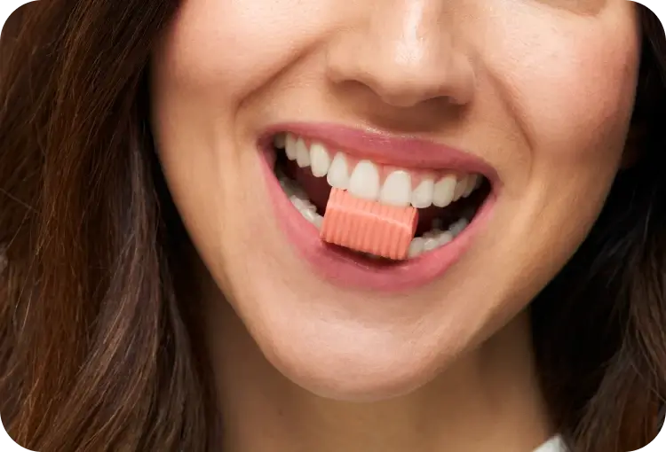A person smiling while holding a pink chewing gum between their teeth.