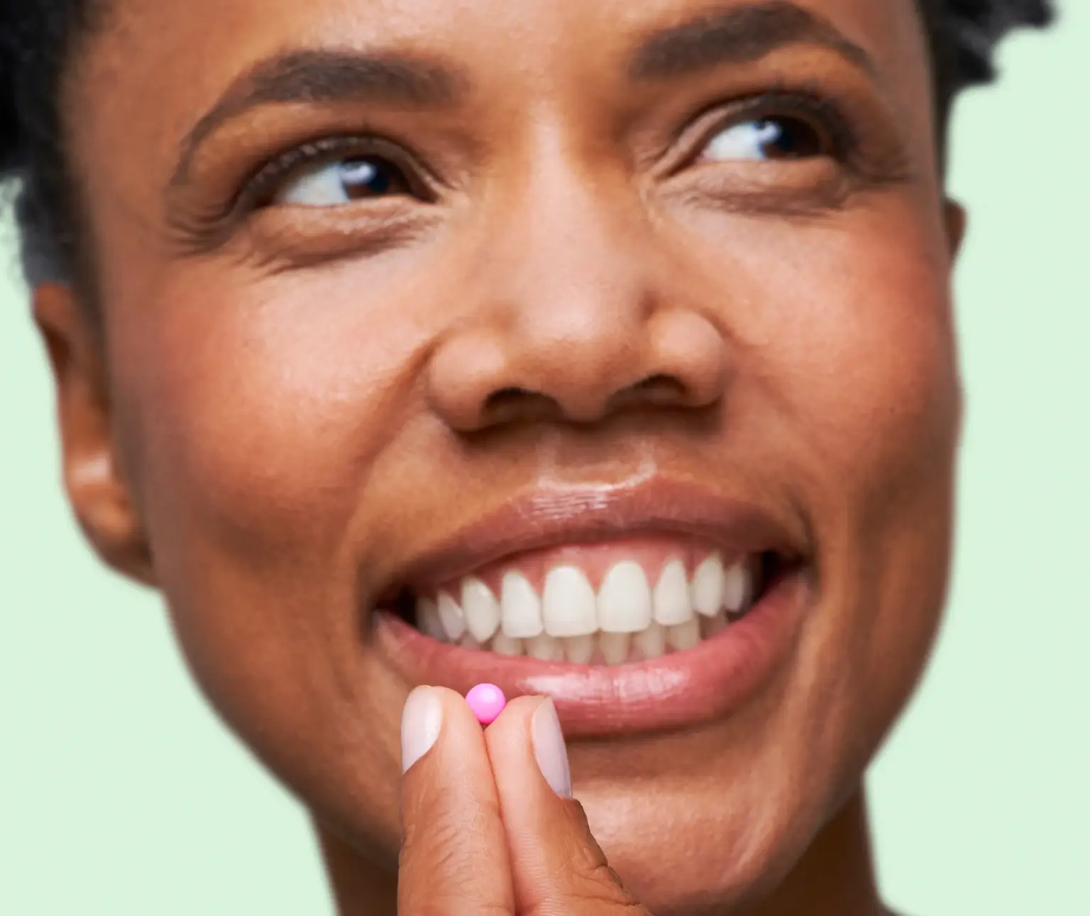 Person smiling while holding a small pink pill near their lips.